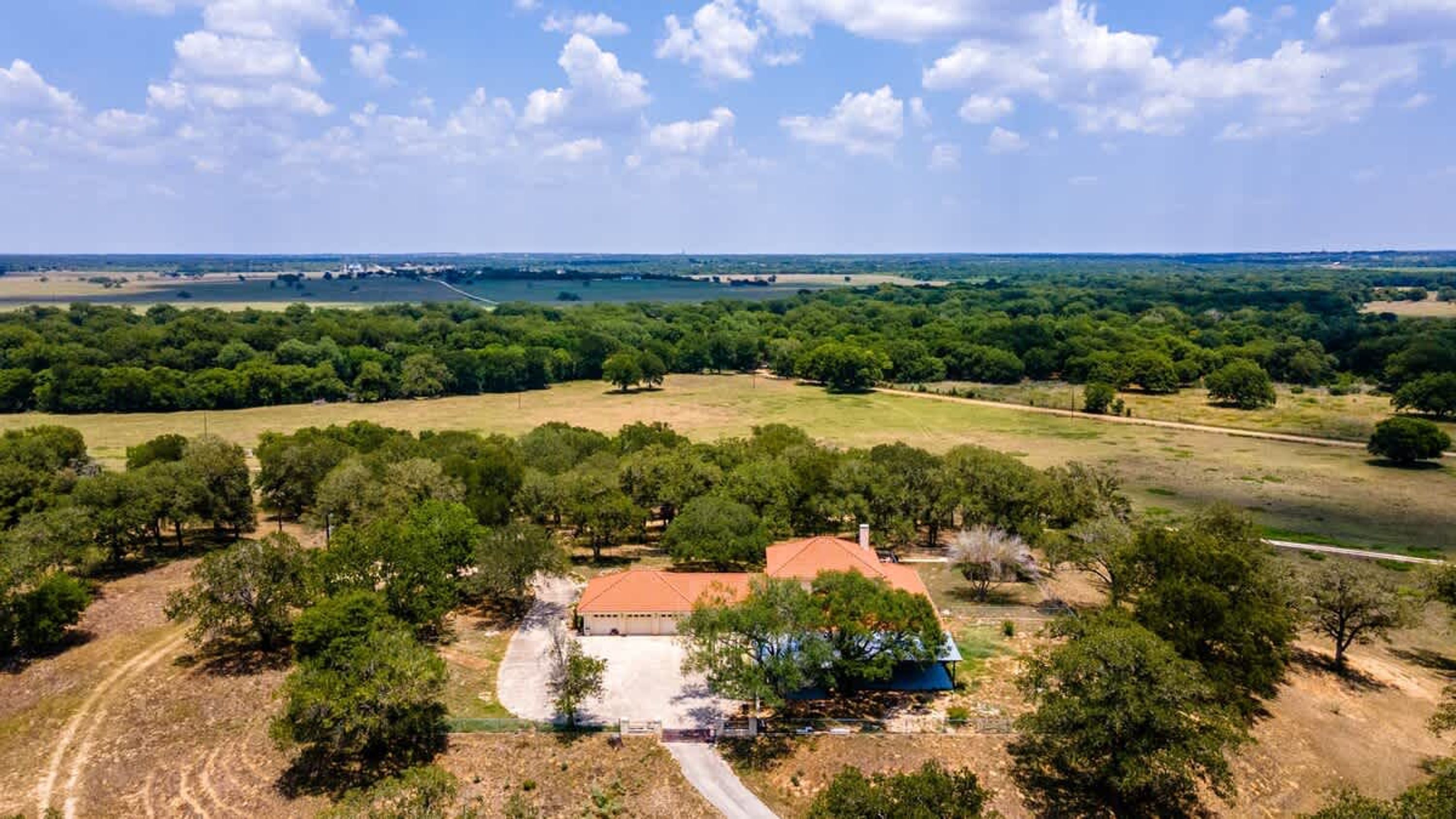 A house surrounded by trees and open fields, with a clear sky and distant water body.