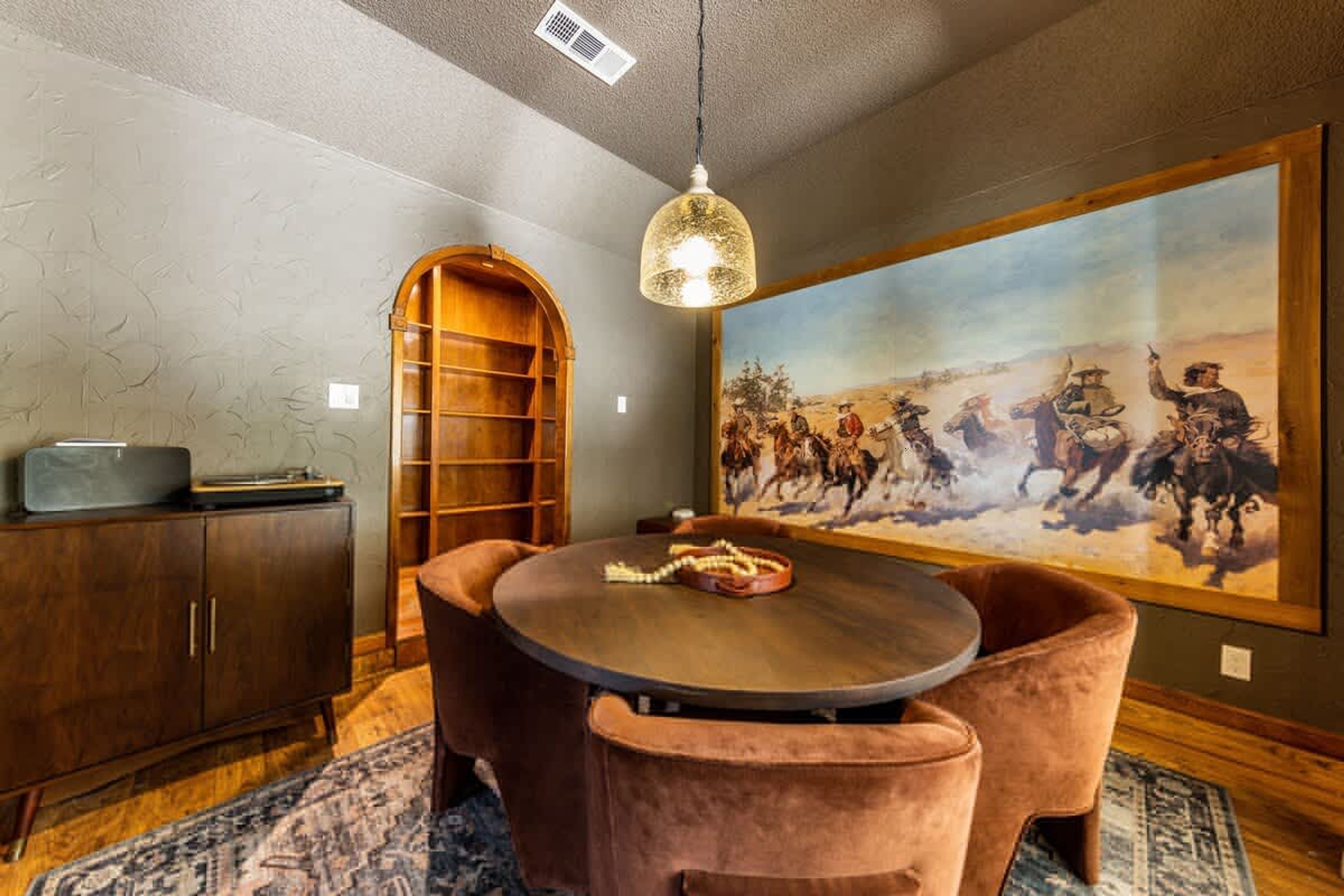 A dining area with a round table, cushioned chairs, a wooden cabinet, a bookshelf, and a large painting on the wall.
