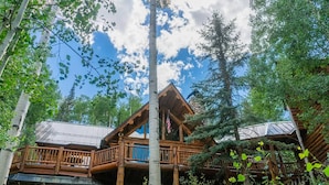 A rustic log cabin with a wooden deck and stone foundation, surrounded by lush greenery and tall trees. - Absolutely Amazing Slopeside Mountain Lodge With spa (Telluride)