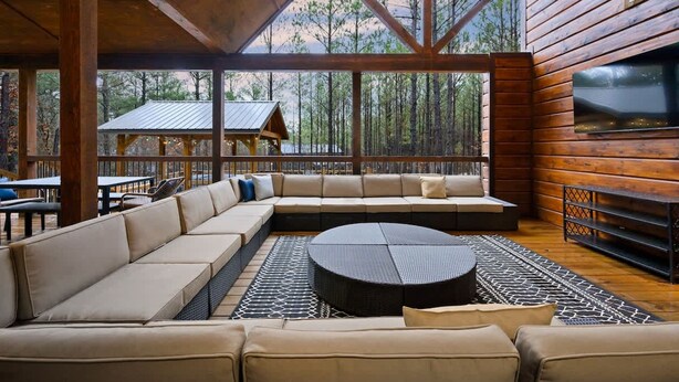 Un vaste espace de détente intérieur, doté d’un grand canapé d’angle et d’une table basse ronde, de grandes fenêtres offrant une vue sur une zone boisée et d’un décor intérieur en bois.