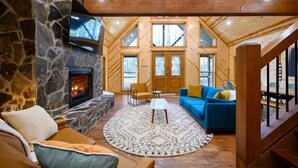 L’intérieur d’un chalet confortable, avec des murs en rondins de bois, une cheminée en pierre, un canapé bleu et une grande fenêtre donnant sur la forêt.