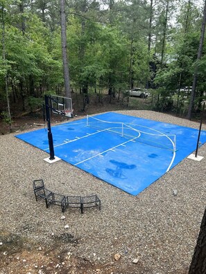 Een buitenbasketbalveld met een blauwe ondergrond, omringd door bomen en grind.