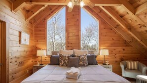 Un dormitorio de cabaña acogedor con cama grande, dos mesitas de noche, una silla y ventana con vistas a árboles.