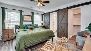 Une chambre à coucher comprenant un lit, une chaise, un bureau, un ventilateur de plafond et une porte coulissante de style rustique.