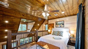 A bedroom with a wooden ceiling, a bed with white bedding, a bench, a nightstand, and a painting on the wall. - Family-friendly Broken Bow Cabin w spa Game Room (Broken Bow)