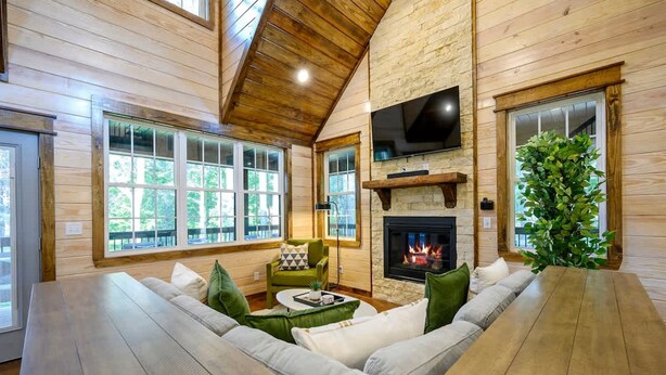 A cozy cabin interior with a fireplace, wooden walls, and a large window overlooking greenery. - Family-friendly Broken Bow Cabin w spa Game Room (Broken Bow)