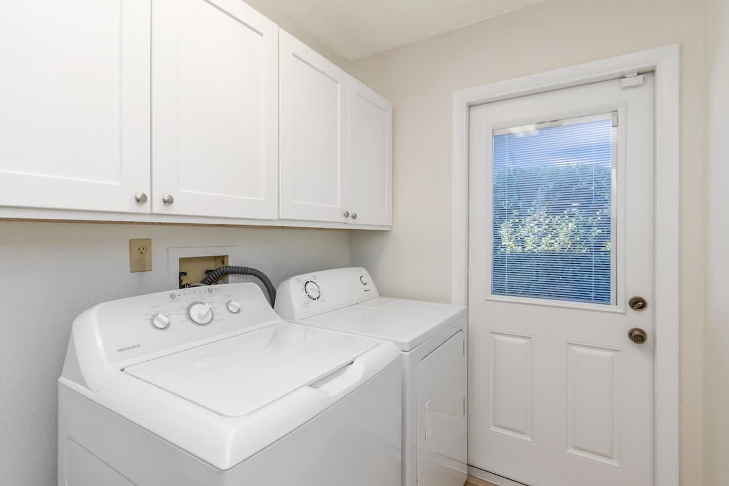 A laundry room with two white front-loading washers and dryers, a white door with a window, and white cabinets above the appliances.