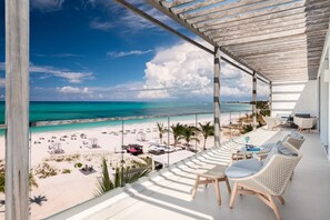 A balcony with a view of a beach and ocean. There are wicker chairs and a table with a drink. - Luxe Oceanfront Penthouse Hot Tub Daily Cabana (Providenciales)