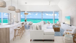 A modern living room with a large window overlooking the ocean, a white sofa, a dining area, and a kitchen with a bar. - Luxe Oceanfront Penthouse Hot Tub Daily Cabana (Providenciales)