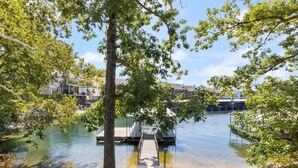 Une vue paisible sur un lac, avec une jetée menant à une plateforme flottante, le tout entouré d’une végétation luxuriante et, au loin, de bâtiments modernes.