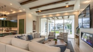 A modern living room with a large sofa, a coffee table, and a fireplace. There is a kitchen area with green cabinets and a dining table. The room has large windows and a view of the outside. - Stunning Vanderbilt Beach House With Heated Pool (Naples)