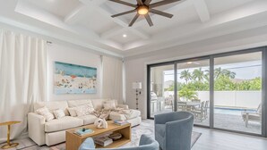 A spacious living room with a white sofa, blue armchairs, a wooden coffee table, and a large window offering a view of a pool and outdoor seating. - Sparkling new Vanderbilt Beach Luxury Escape (Naples)