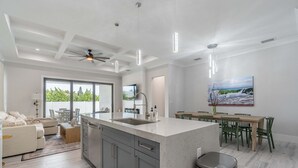 A modern kitchen with a large island, a dining area with green chairs, and a view of greenery outside. - Sparkling new Vanderbilt Beach Luxury Escape (Naples)