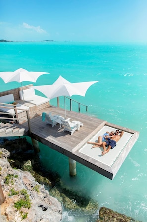 A couple relaxing on a wooden deck with white lounge chairs and umbrellas, overlooking turquoise waters.