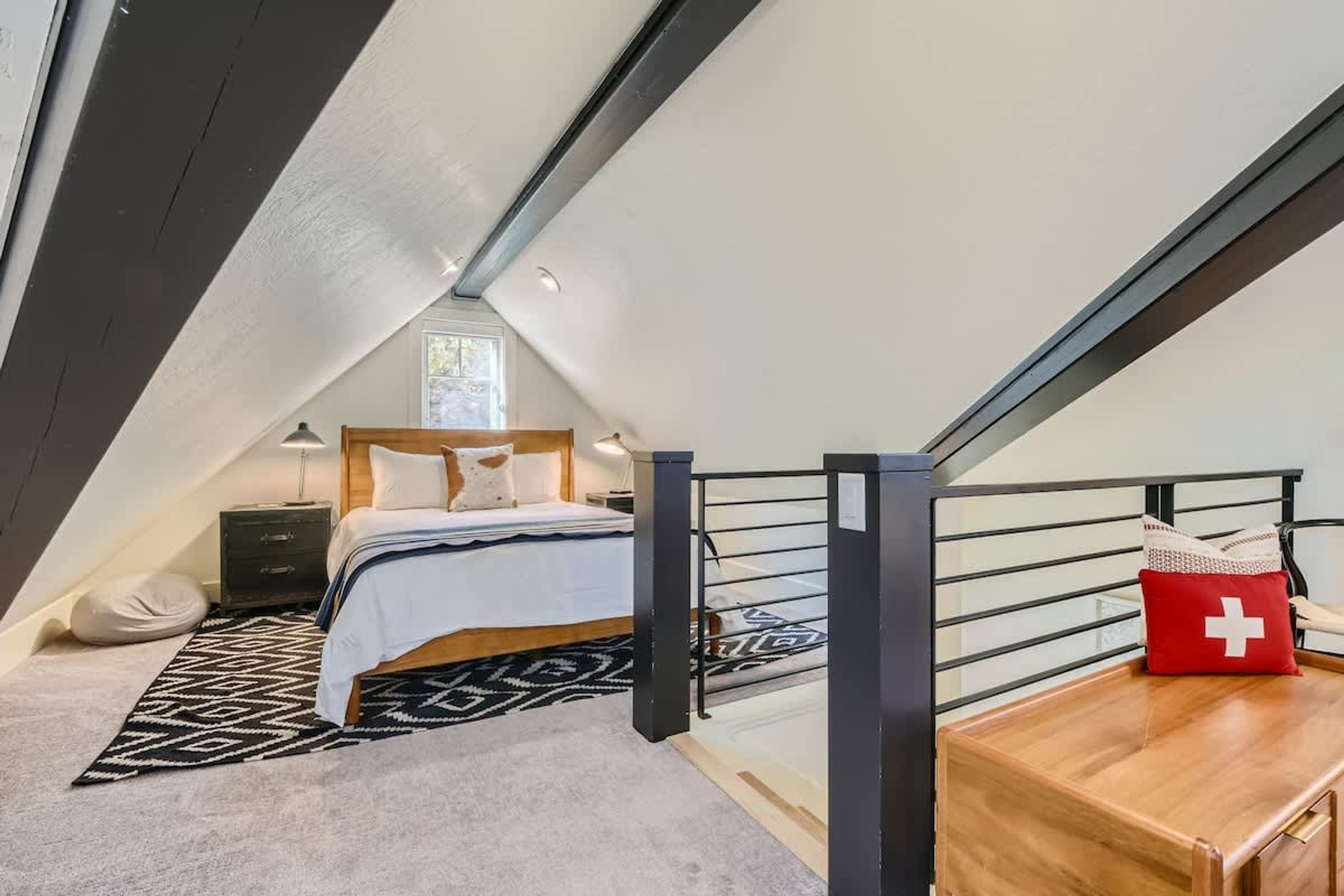 A bedroom with a bed, bedside table, lamp, and a red cushion with a white cross.