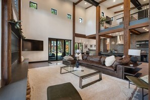 A spacious living area with a large leather sofa, a glass coffee table, and a fireplace. There is a kitchen visible in the background with a brick wall and modern appliances. - Architecturally Stunning Cle Elum Luxury Retreat (Cle Elum)