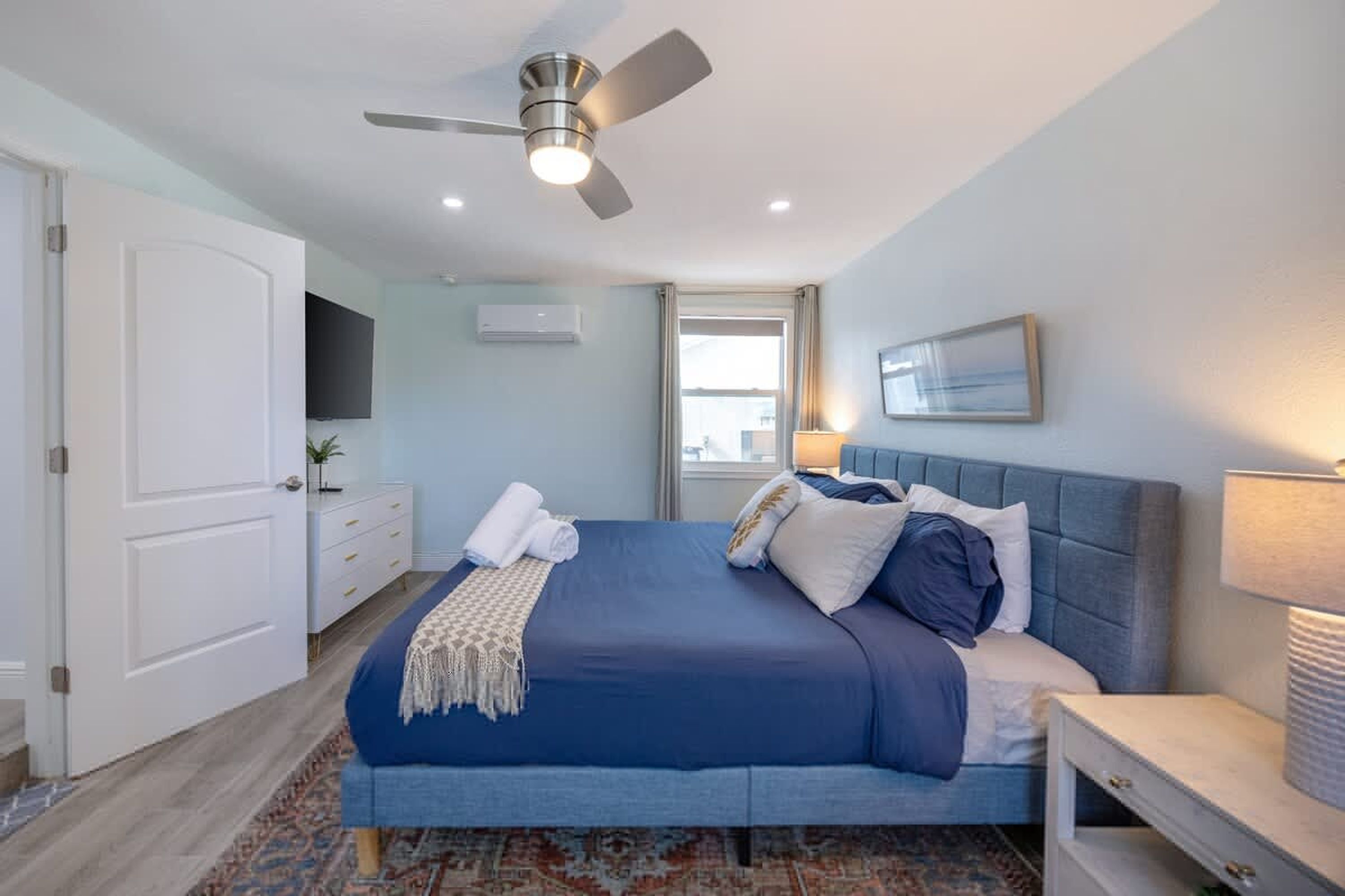 A bedroom with a bed, a ceiling fan, a bedside table, a window, and a door.