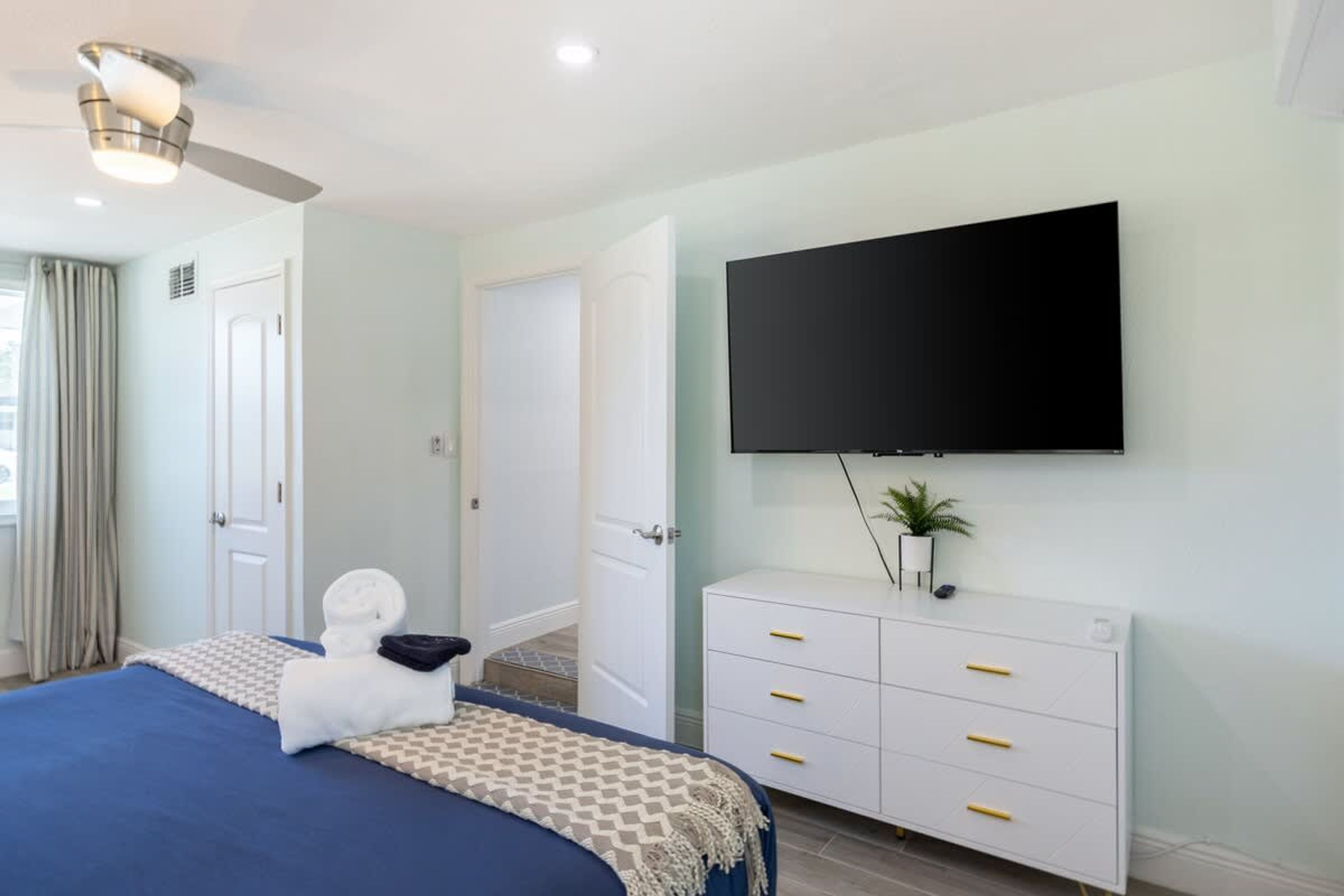 A hotel room with a bed, a TV mounted on the wall, a white dresser, and a ceiling fan.