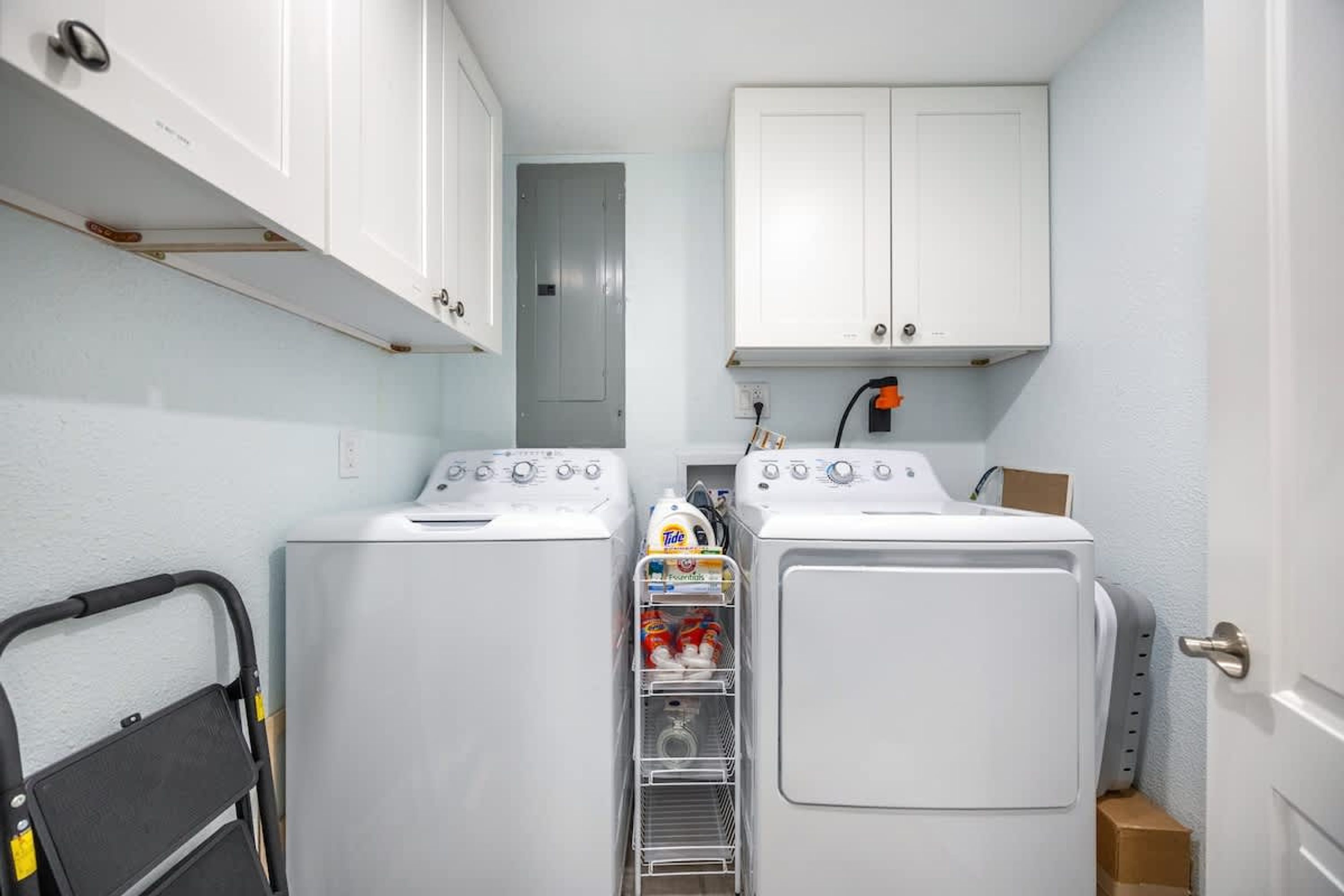 A compact laundry room with two front-loading washing machines, a dryer, and a small storage shelf with laundry supplies.