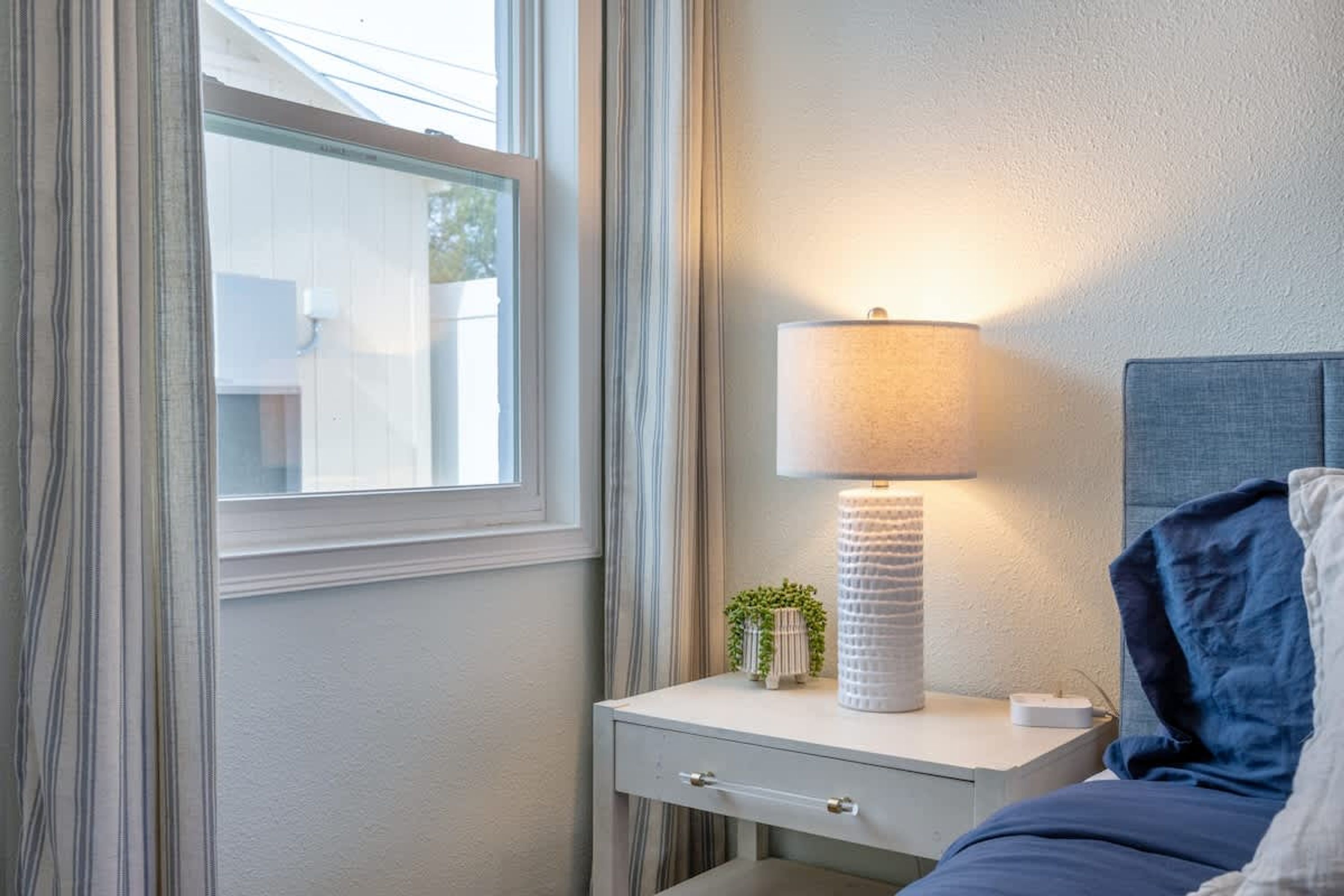 A neatly arranged bedroom with a bedside table, a lamp, a small plant, and a bed with a blue blanket.