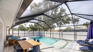 A covered outdoor pool area with a wooden table and chairs, lounge chairs, and a view of trees and a fence in the background. - Fun Family-friendly Largo Home Heated Pool (Largo)
