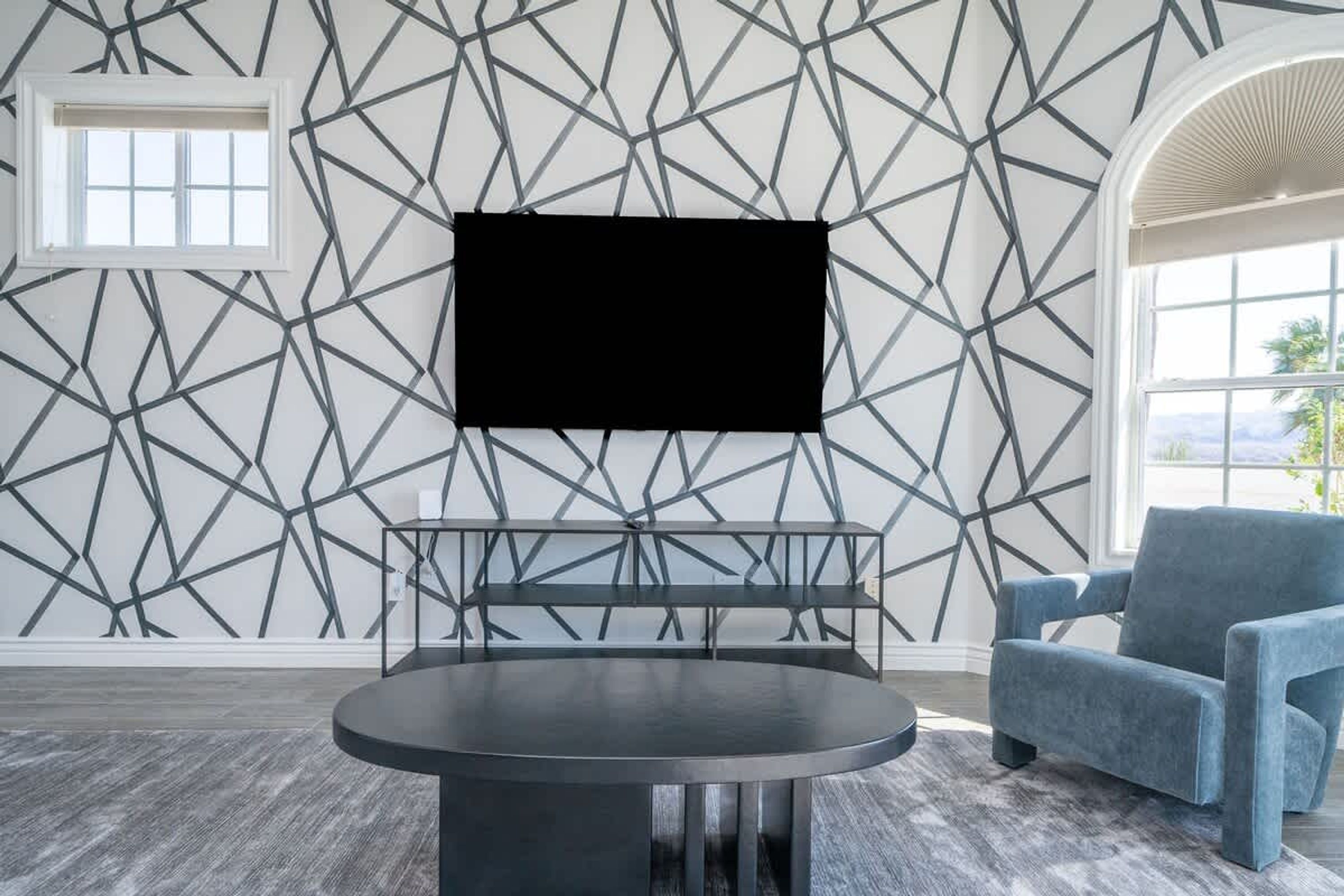 A room with a geometric-patterned wall, a round table, a blue armchair, and a window with a view.