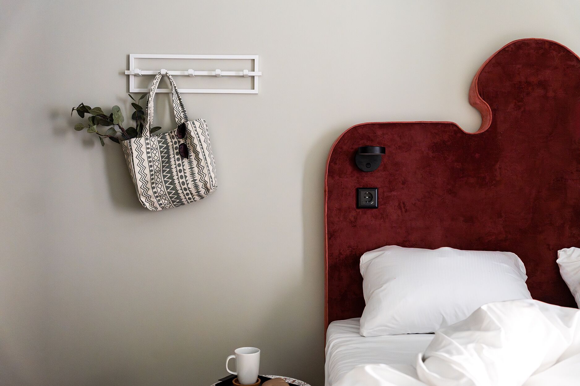 A hotel room with a bed, a bedside table with a cup, a wall-mounted light, and a hanging bag.