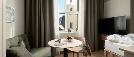 Quarto de hotel com cama, televisão, escrivaninha com cadeira, mesinha com um recipiente de lanches e um sofá.