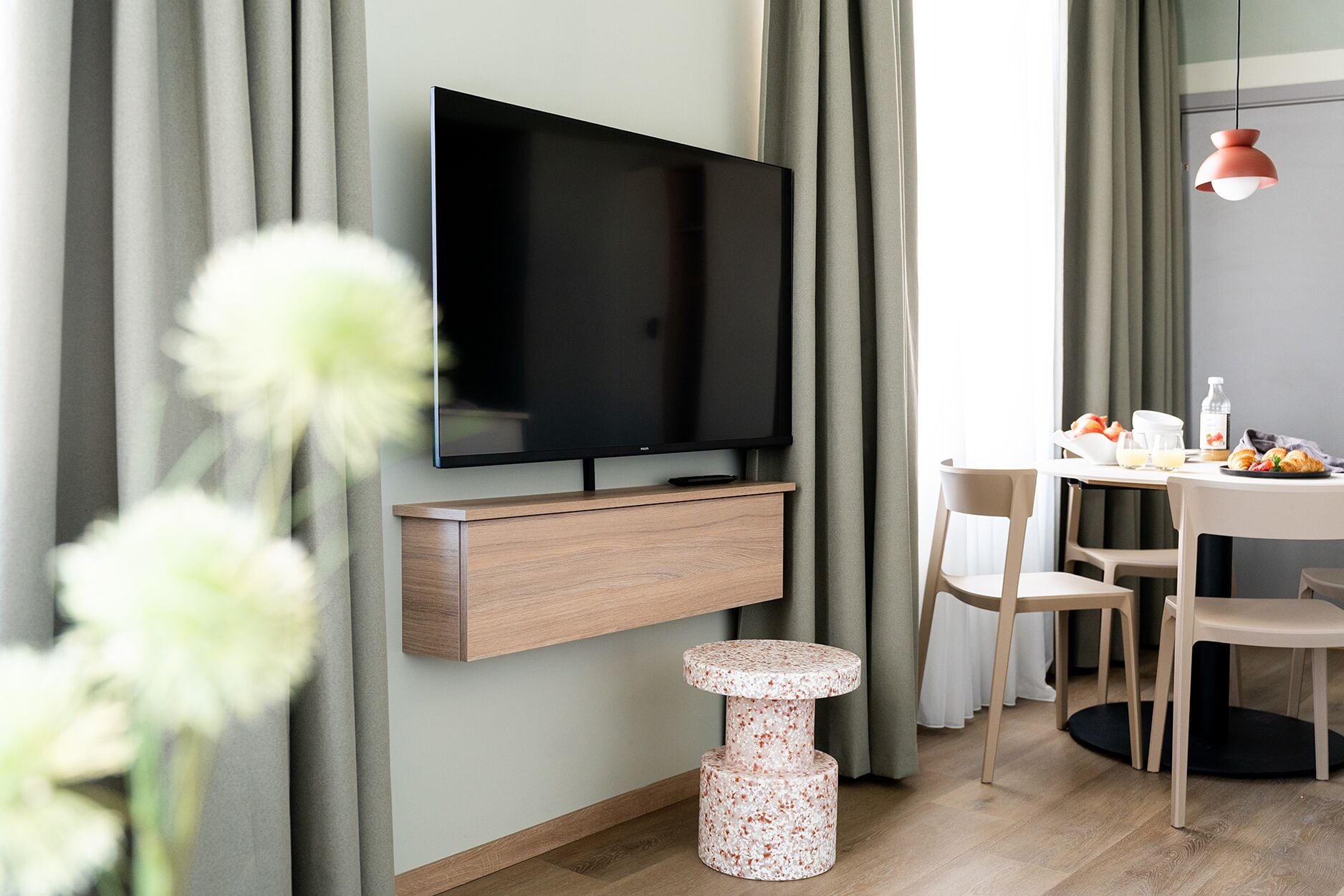 Un soggiorno moderno con una TV a schermo piatto montata a parete, un mobiletto in legno sottostante, una cuccia rotonda per animali domestici e un'area pranzo con sedie e un tavolo.