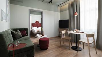 Et moderne hotelværelse med en grøn sofa, et lille rundt bord med stole, en seng med rødt sengegavl, et fjernsyn og et stort vindue med gardiner.