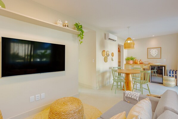A modern living room with a flat-screen TV, a sofa, a dining area with a table and chairs, and a kitchen area with a refrigerator and a sink.