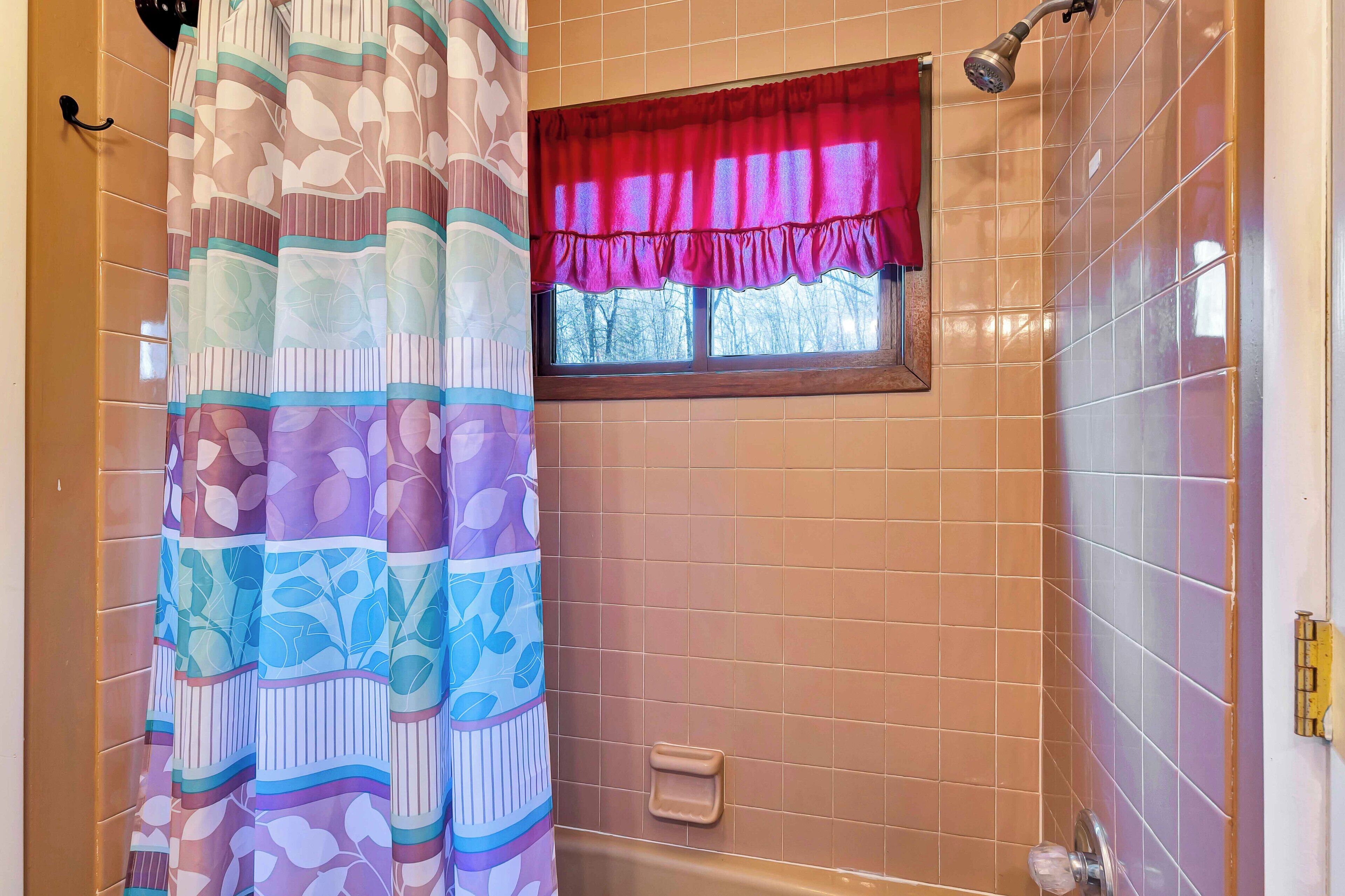 A bathroom with a shower, a window with a red valance, and a patterned shower curtain.