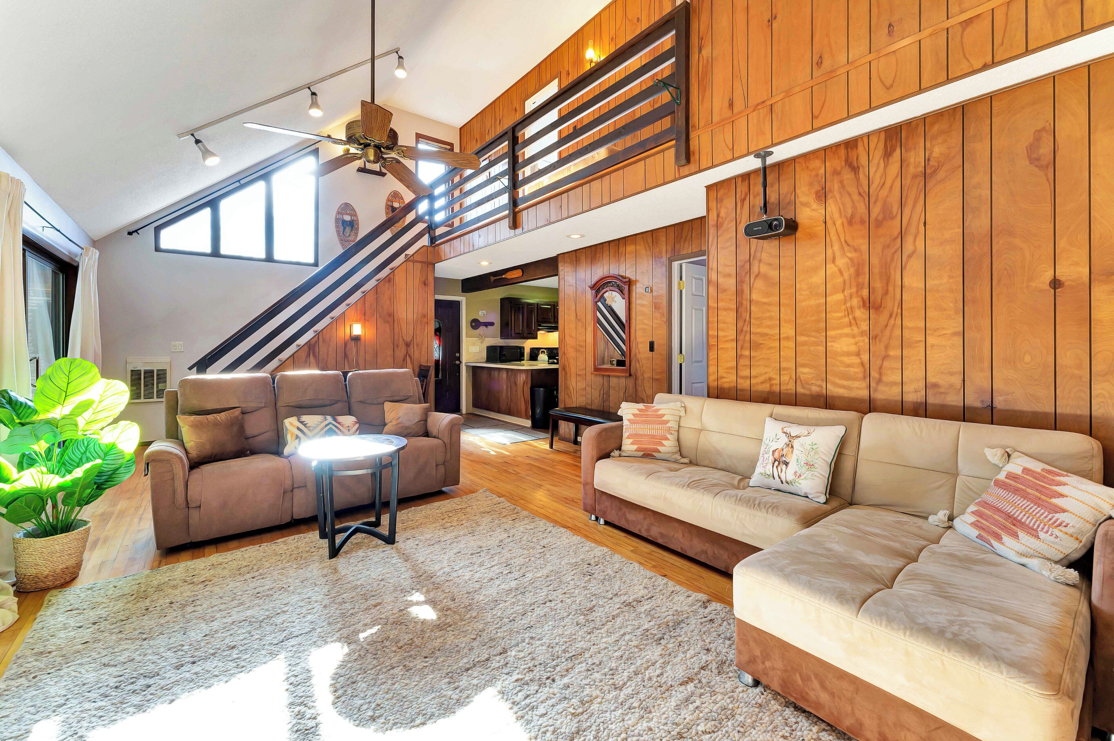 A spacious living area with a high ceiling, wooden paneling, a staircase, a sofa set, and a large plant.