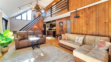 A spacious living area with a high ceiling, wooden paneling, a staircase, a sofa set, and a large plant.