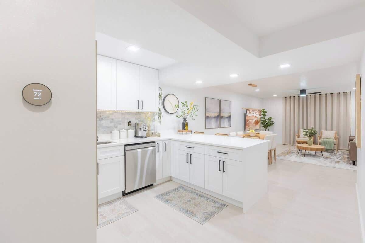 A modern kitchen with white cabinetry, a stainless steel dishwasher, and a central island. There is a clock on the wall and a plant on the island.