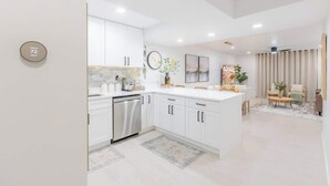 Una cocina moderna con armarios blancos, lavavajillas de acero inoxidable y una isla central. Hay un reloj en la pared y una planta en la isla.