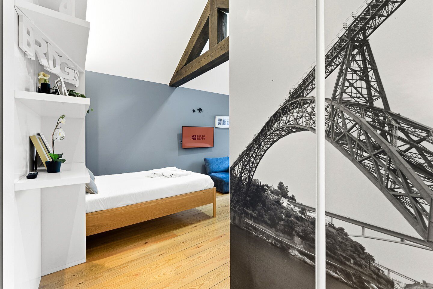 A room with a bed, a shelf with books and a plant, a 'GUEST READY' sign, and a wall with a black and white photo of a bridge.