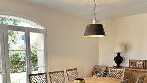 A dining area with a round wooden table, wicker chairs, a hanging light fixture, and a view through glass doors. - Les Lauriers Roses house with swimming pool - popular area Isle sur la Sorgue (L'Isle-sur-la-Sorgue)