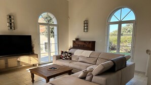 A spacious living room with a large sofa, a wooden coffee table, and a flat-screen TV. There are two large windows providing natural light and a view of the outdoors. - Les Lauriers Roses house with swimming pool - popular area Isle sur la Sorgue (L'Isle-sur-la-Sorgue)