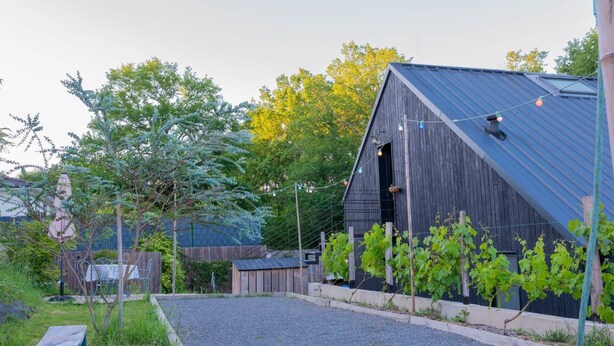 A rustic barn with a sloped roof, a gravel driveway, and a vineyard in the foreground. - Studio Mandavi - Vineyards & Forest (Loupes)