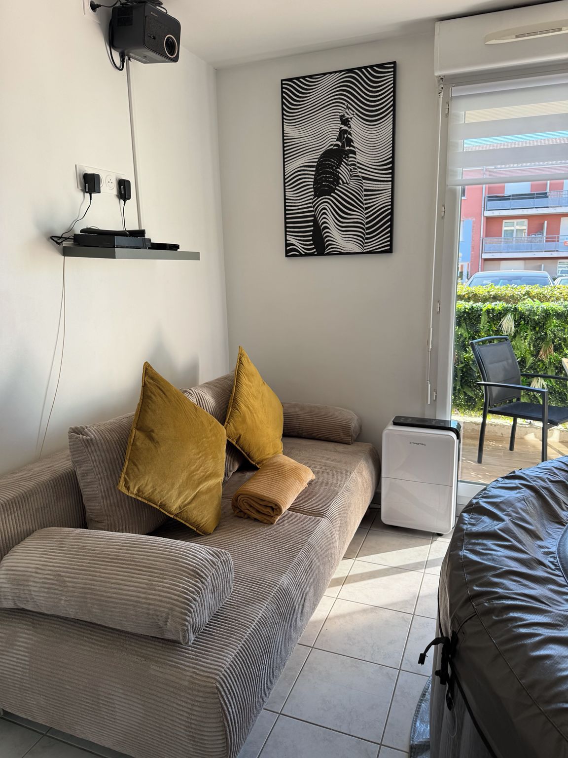 A modern living room with a sofa, a projector, a heater, and a window with a view of buildings and greenery.