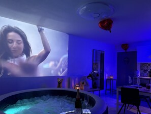 A modern room with a large screen displaying a movie, a hot tub, and a dining area with a table set for two. - Suite Eden Spa, Appart’hôtel bien être , Patio (Avion)