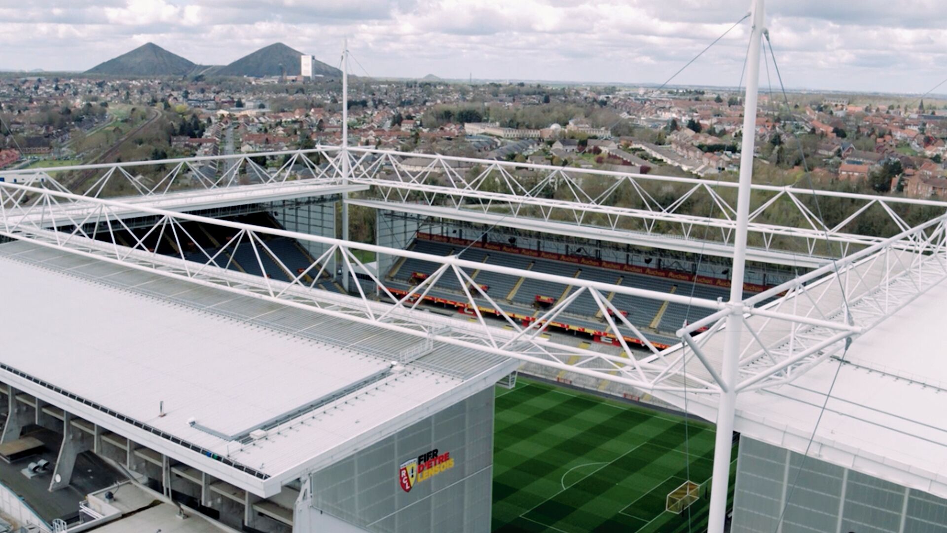 A stadium with a white roof and a green field, surrounded by a cityscape and mountains.