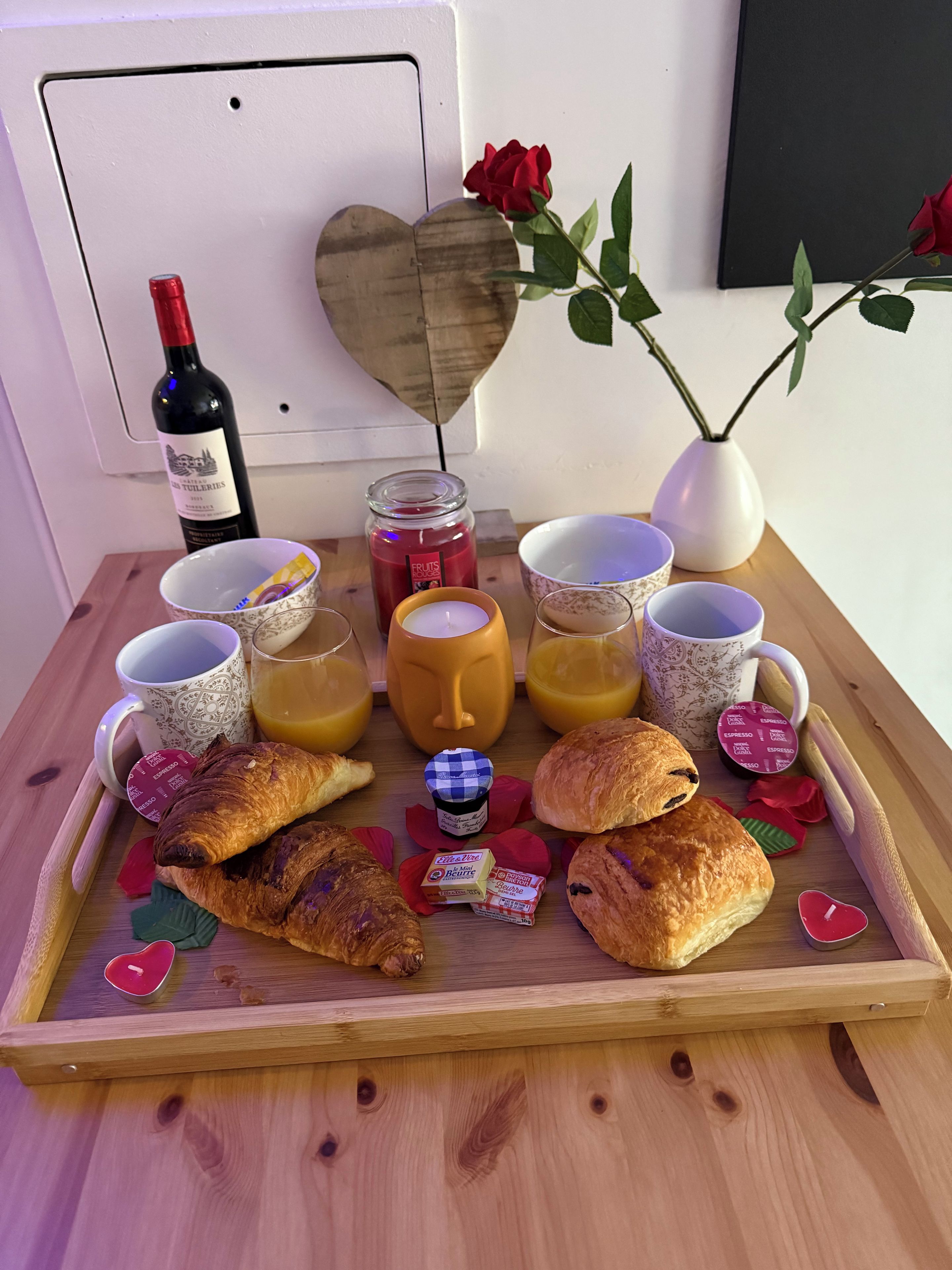 A breakfast tray with croissants, a bottle of wine, a candle, and a vase with a rose.