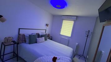 Un dormitorio con cama, mesitas de noche, una ventana con persianas, una lámpara de techo y un televisor montado en la pared.