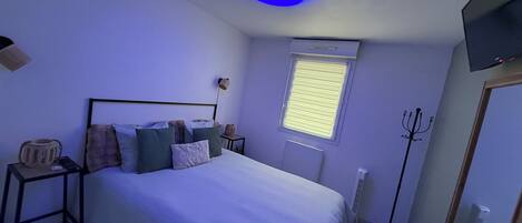 Une chambre à coucher comprenant un lit, des tables de chevet, une fenêtre avec des stores, un luminaire au plafond et un téléviseur fixé au mur.