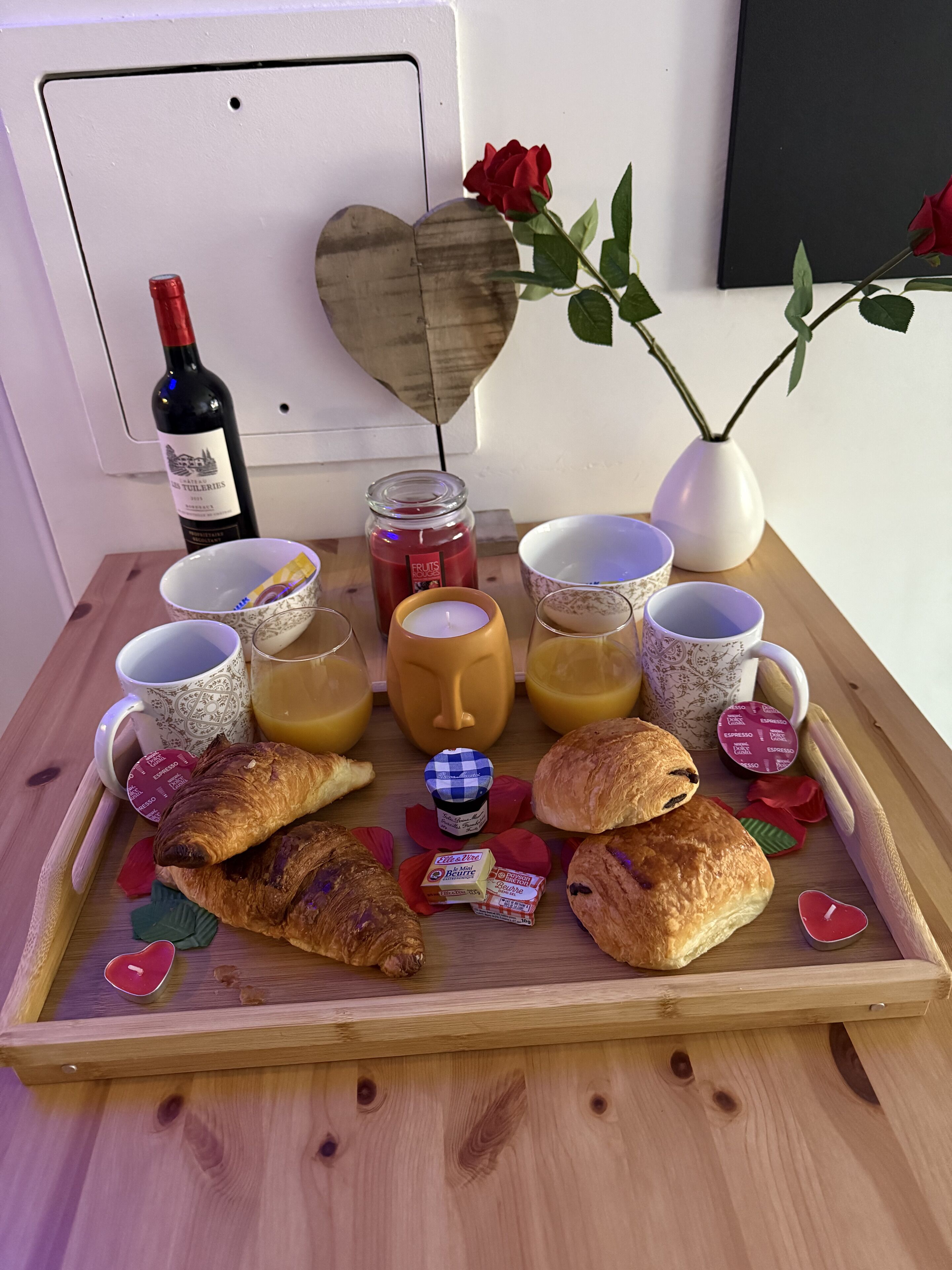 Un vassoio di legno con prodotti per la colazione, tra cui croissant, una candela a forma di cuore, una bottiglia di vino e un vaso con una rosa rossa.