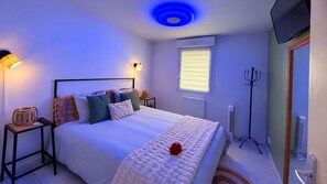 A bedroom with a bed, bedside tables, a lamp, a heater, a TV, and a window with blinds. - Suite Eden Spa, Appart’hôtel bien être , Patio (Avion)