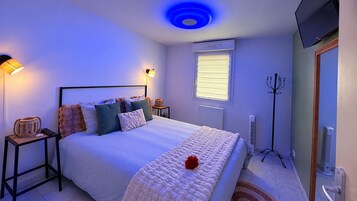 Un dormitorio con cama, mesitas de noche, lámpara, calefactor, televisor y ventana con persianas.