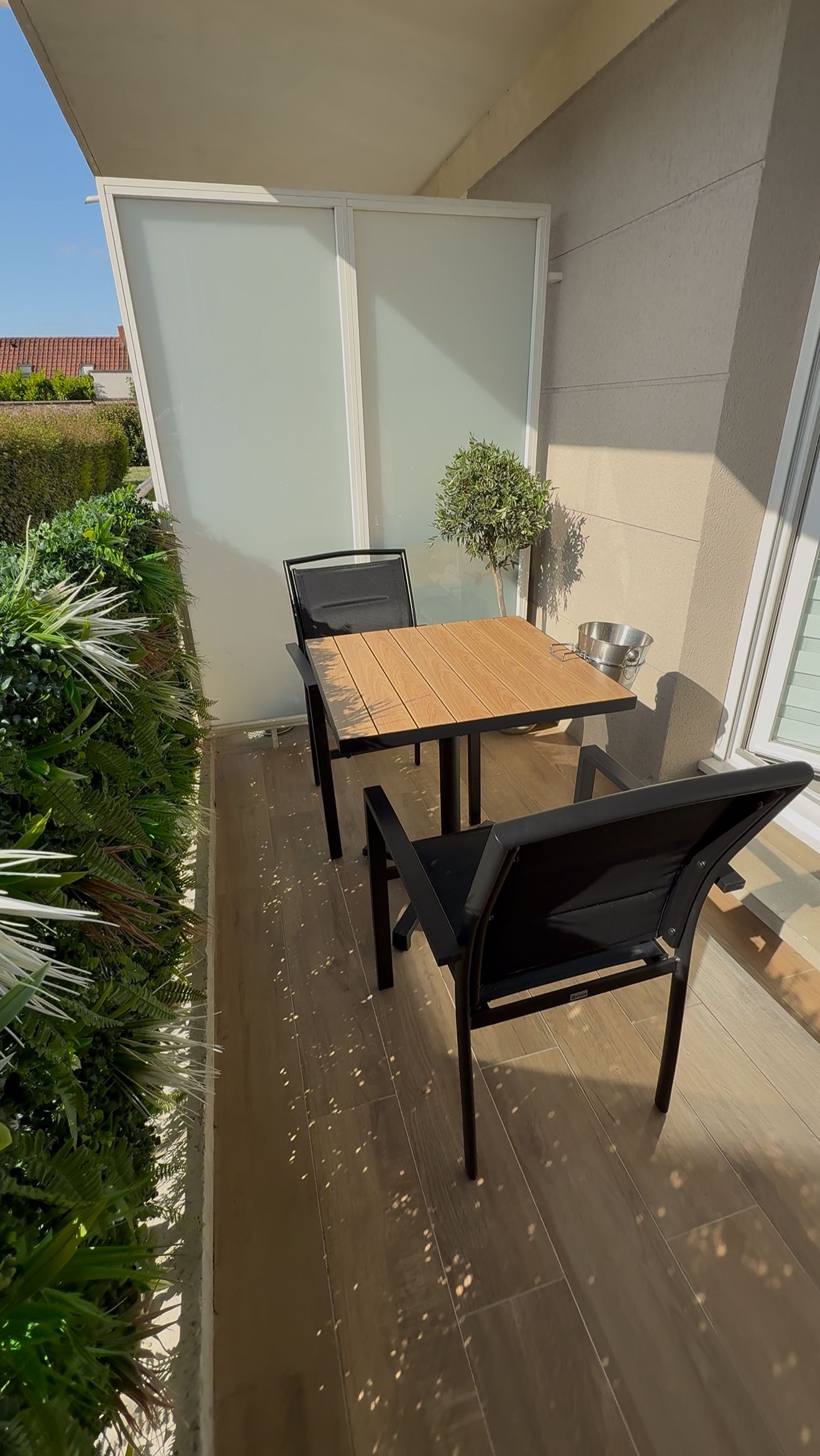 Une terrasse extérieure avec une table en bois et des chaises noires, une petite plante en pot et un seau en métal.